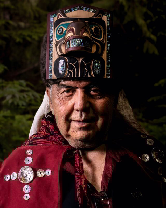 An elder wearing a traditional robe decorated with decorative buttons and a carved and painted wooden headdress stands in front of a background of blurred leaves.