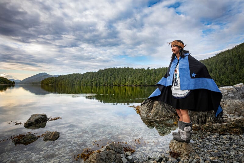 A person in traditional indigenous costume stands on a rock by a quiet lake surrounded by forested hills and a cloudy sky, with mountains visible in the distance.