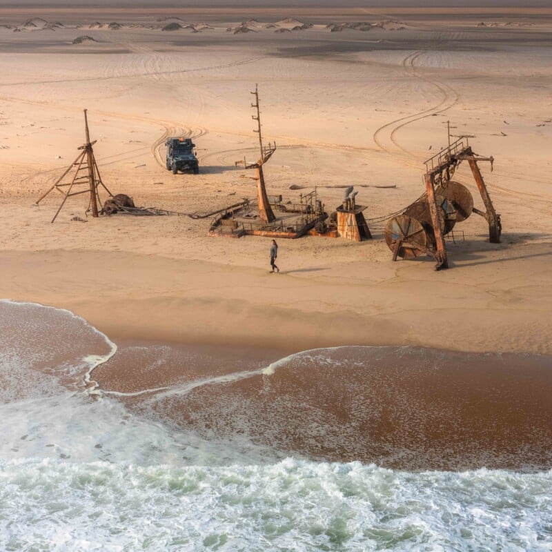 A person walks near the rusted remains of a shipwreck on a sandy beach. You can see ocean waves in the foreground and a parked car in the background of sand dunes.
