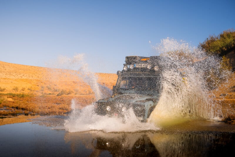 A rugged off-road vehicle drives through shallow water streams in a desert landscape, making big splashes. Sunlit dunes and clear blue sky are visible in the background.