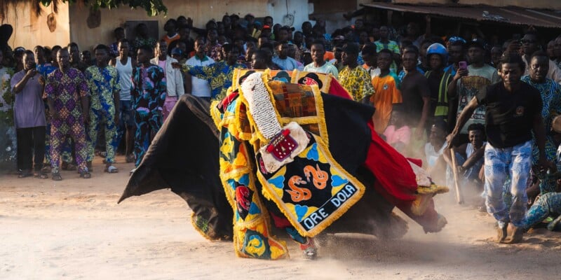 Performers in colorful beaded masquerade costumes dance energetically in the center of a dusty outdoor area, surrounded by onlookers, many wearing brightly patterned clothing.