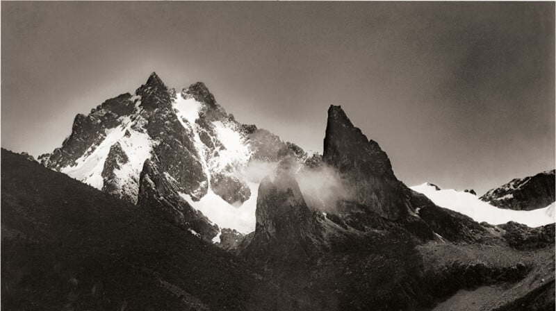 Snow-capped mountain peaks with rugged, jagged edges rise sharply against the cloudy sky. Fog and clouds drift around the rocky peaks, highlighting the dramatic and contrasting landscape.