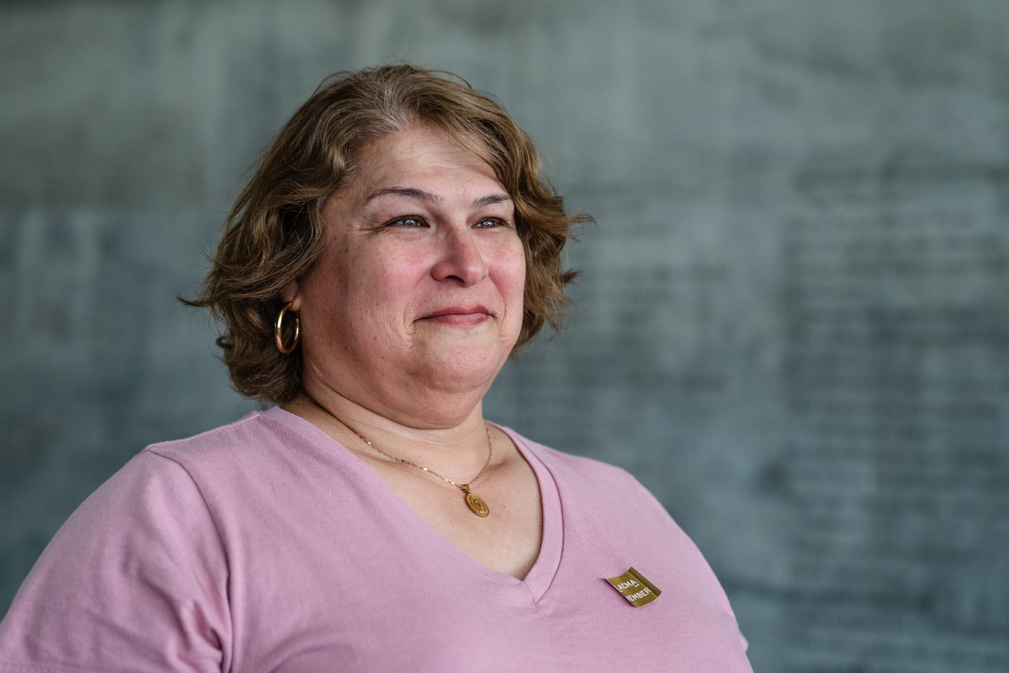 A woman in a pink shirt stands in front of a concrete wall in a museum. 