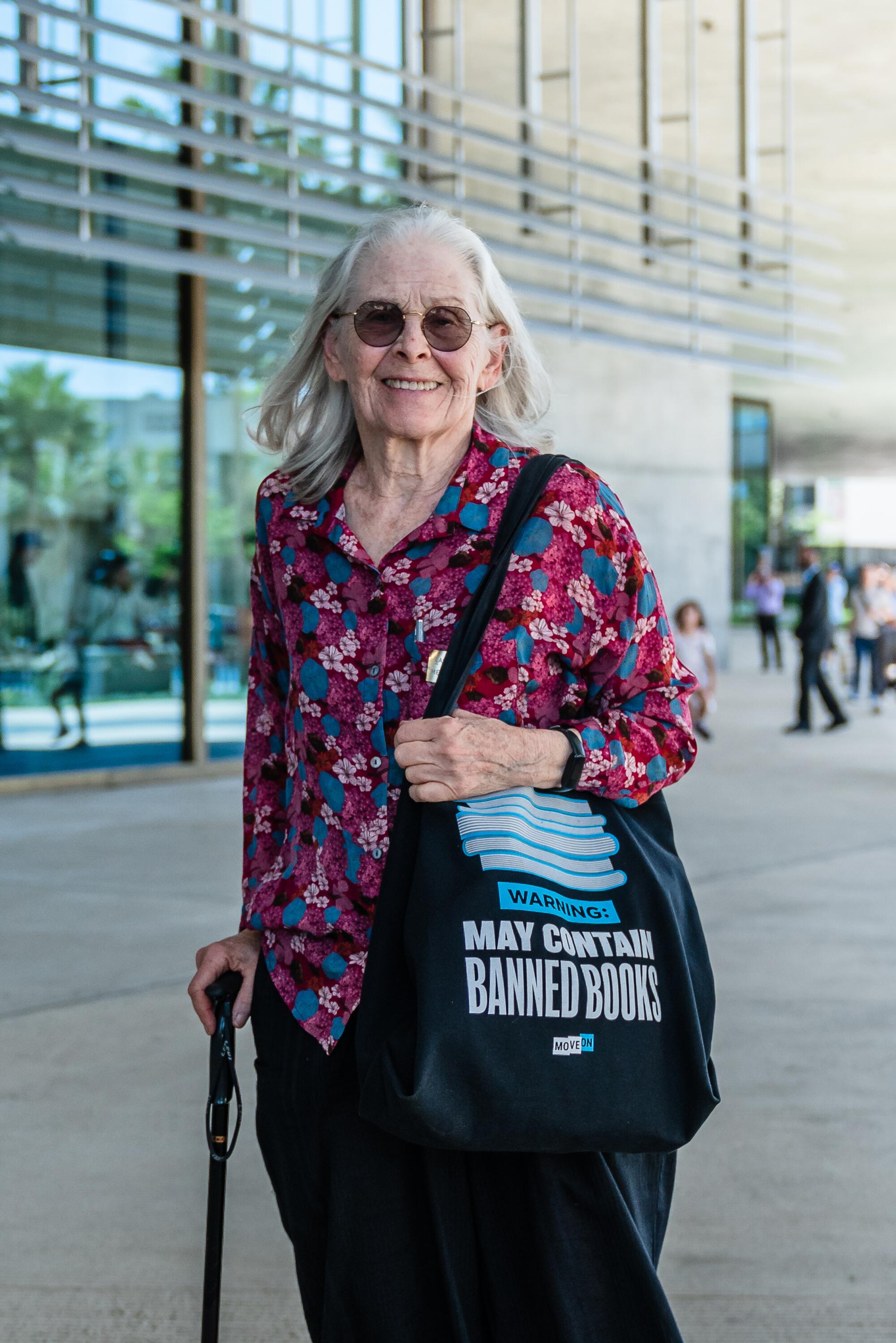 A woman with a cane and a tote bag stands in front of a museum.